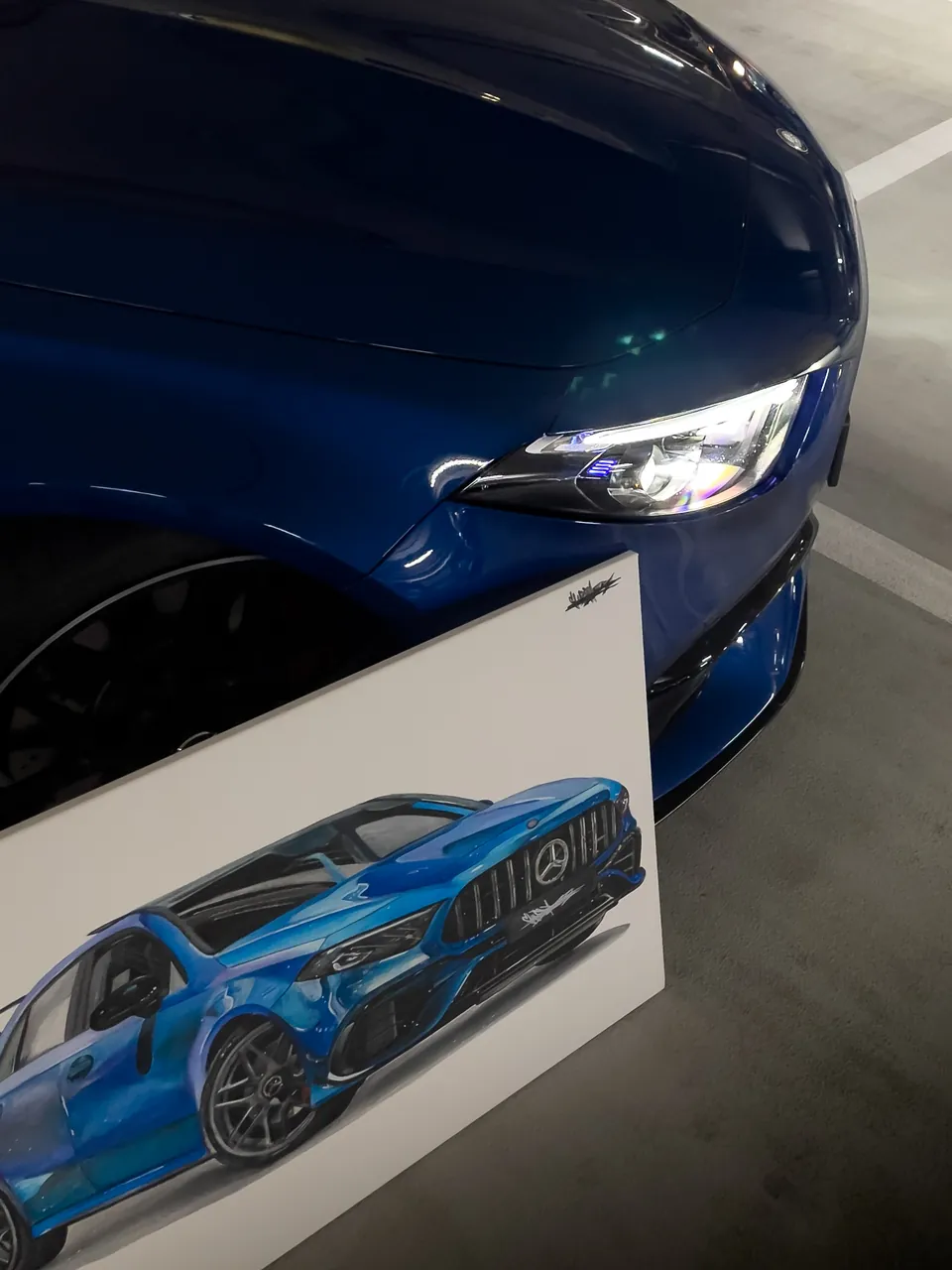 Obraz modrého Mercedesu-AMG opřený o kolo skutečného vozu, detail světla